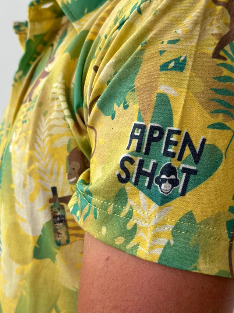 Een close-up van een persoon die een geel shirt met een opdruk van bladeren en de tekst 'APEN SH0T' draagt.